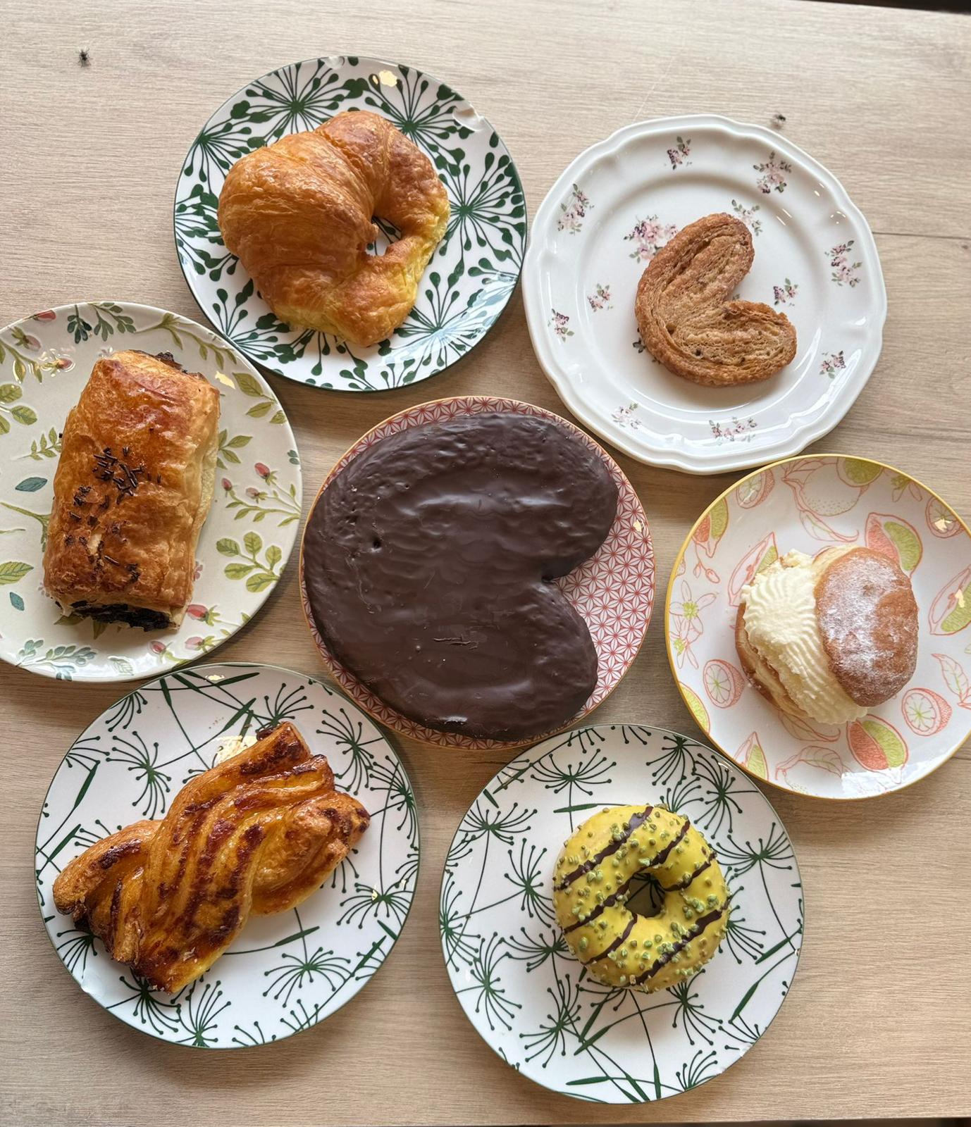 Bollería surtida vista cenital en platos florales: croissant, napolitana de chocolate, donut glaseado de limón, palmera y bollo de nata
