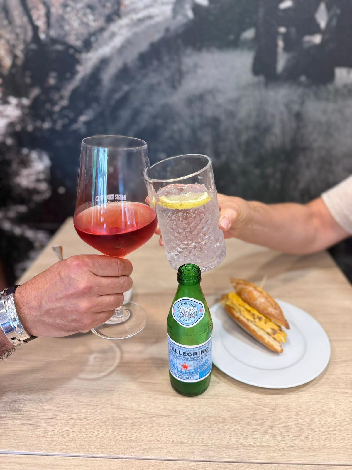 Brindis con copa de vino rosado y agua con limón junto a bocadillo de tortilla sobre mesa de madera en Sara Kafetegia