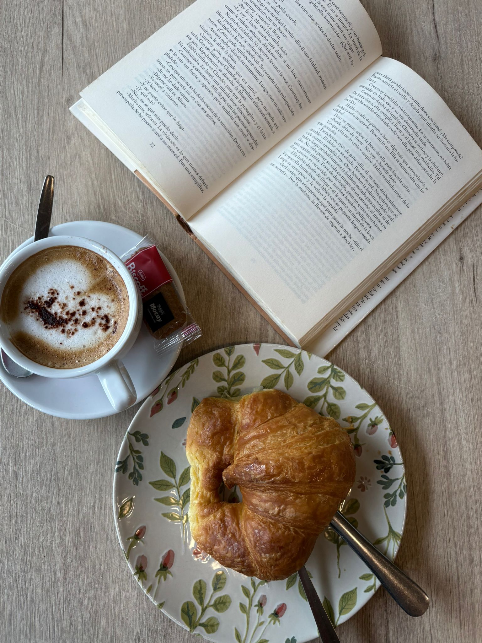 Cappuccino cenital con latte art de corazón y cacao, junto a croissant dorado en plato floral y libro abierto