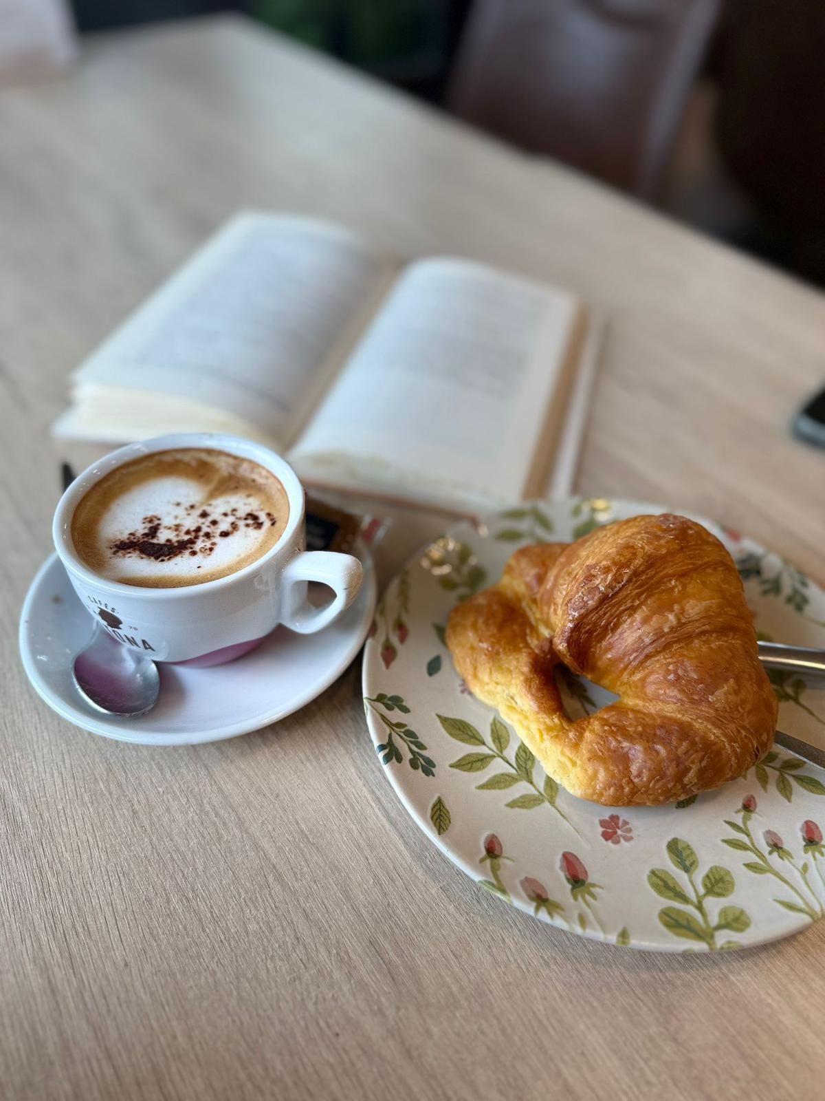 Cappuccino con latte art de corazón espolvoreado de cacao y croissant dorado en plato floral sobre libro abierto, desayuno en Sara Kafetegia