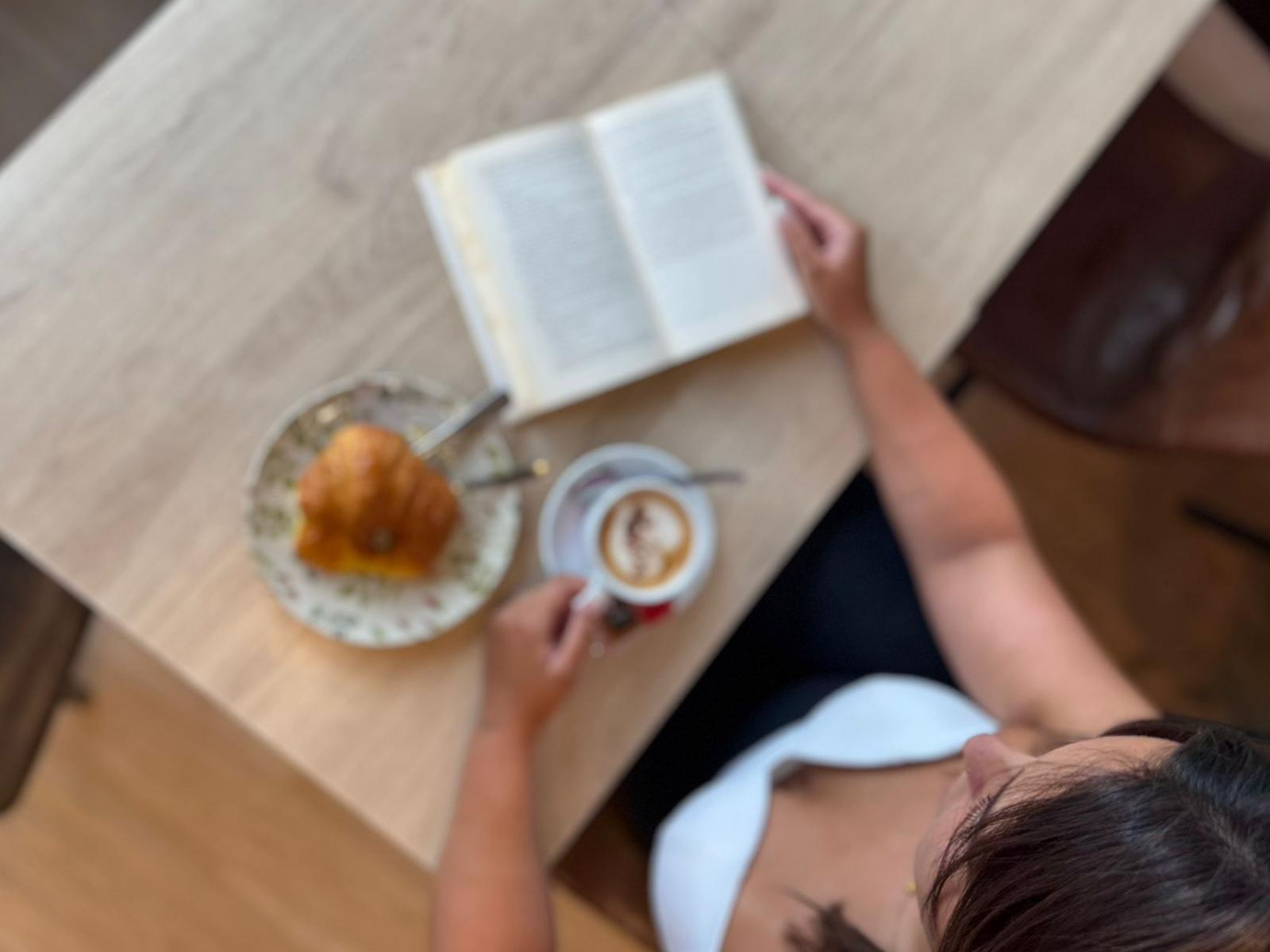 Cappuccino con latte art de corazón y cacao junto a croissant dorado en plato floral y libro abierto, fotografía cenital en Sara Kafetegia