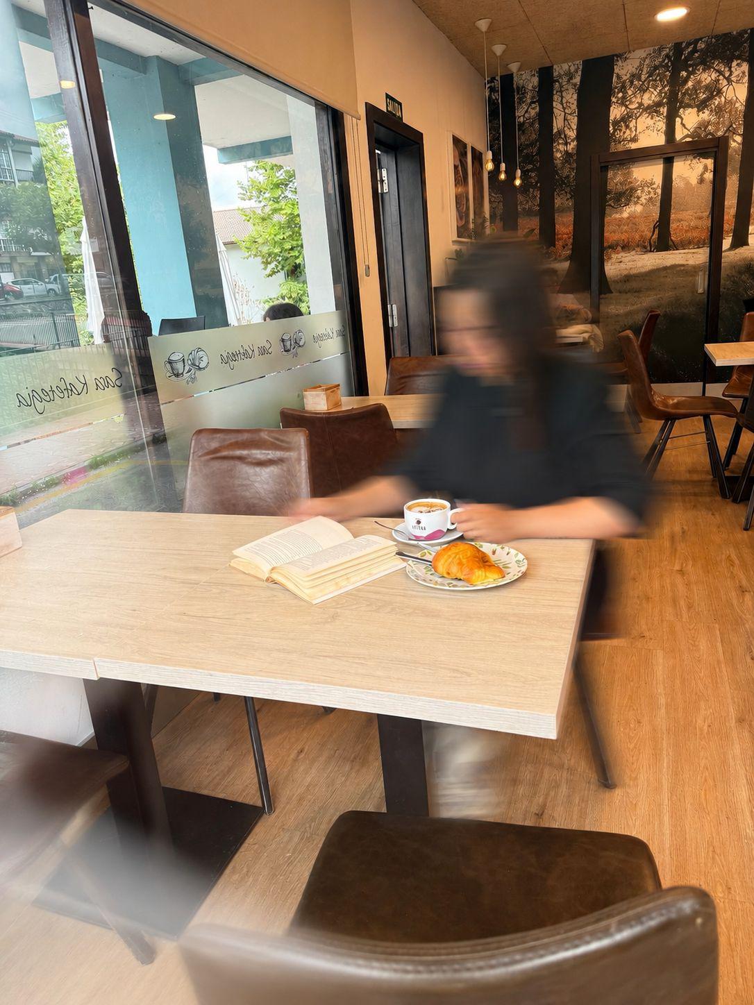 Cliente leyendo con un café y croissant en mesa de Sara Kafetegia, con el mural de bosque y las bombillas Edison al fondo