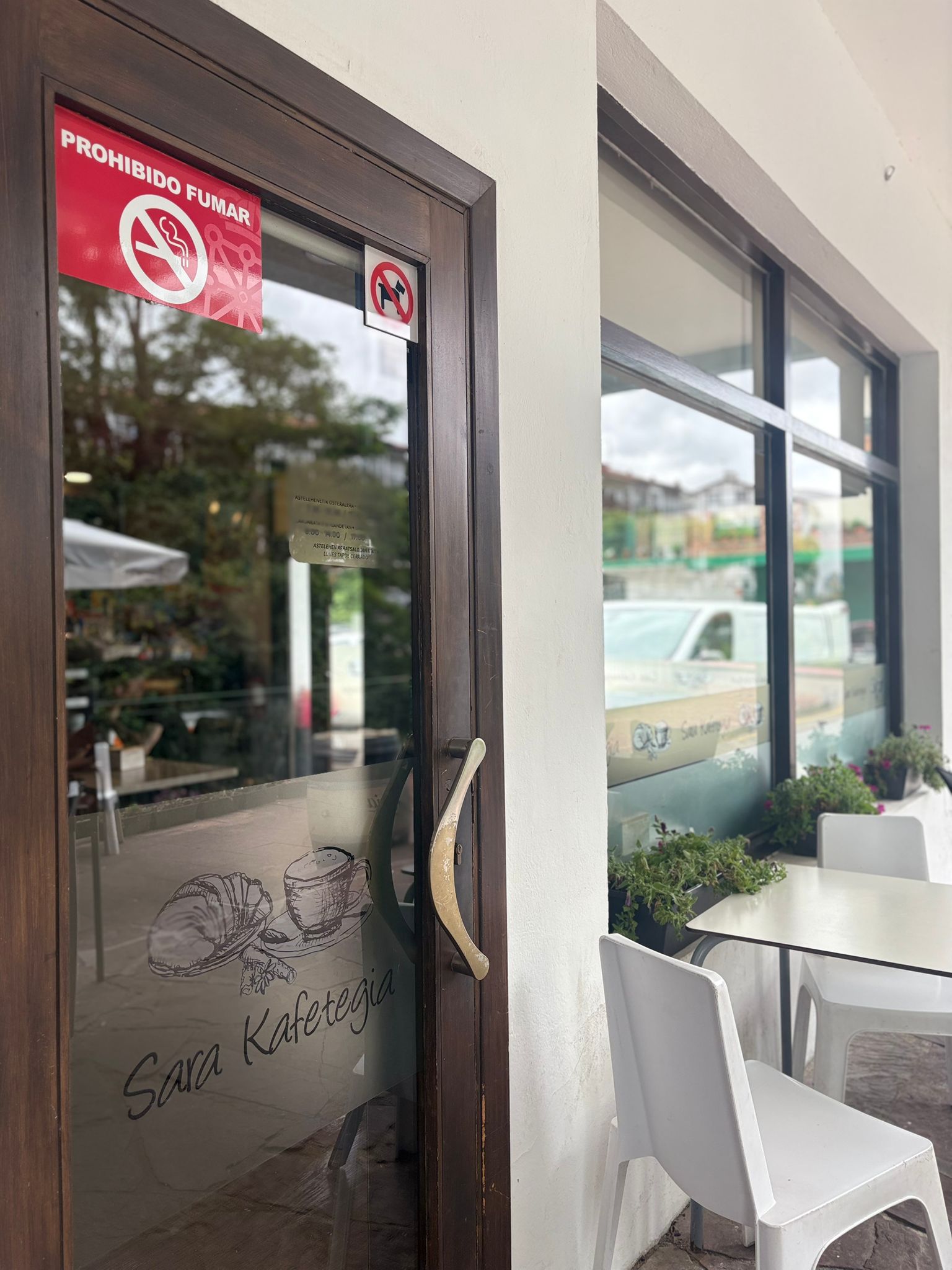Puerta de entrada de Sara Kafetegia con cristal y logo grabado, terraza con sillas blancas y flores en la fachada