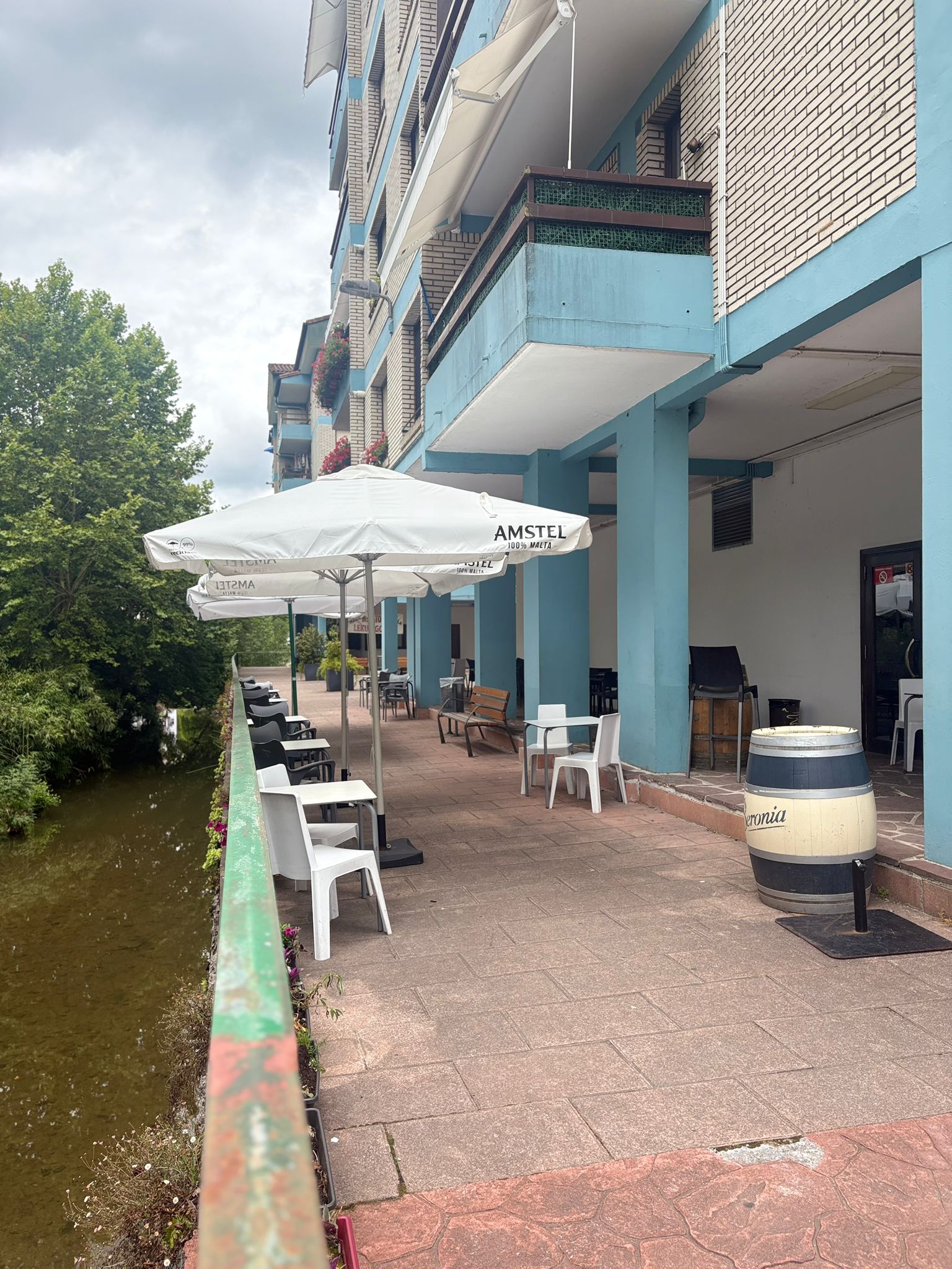 Terraza larga de Sara Kafetegia con pórtico de pilares azules, sombrillas Amstel, mesas blancas y río Bidasoa a la izquierda entre arboleda verde