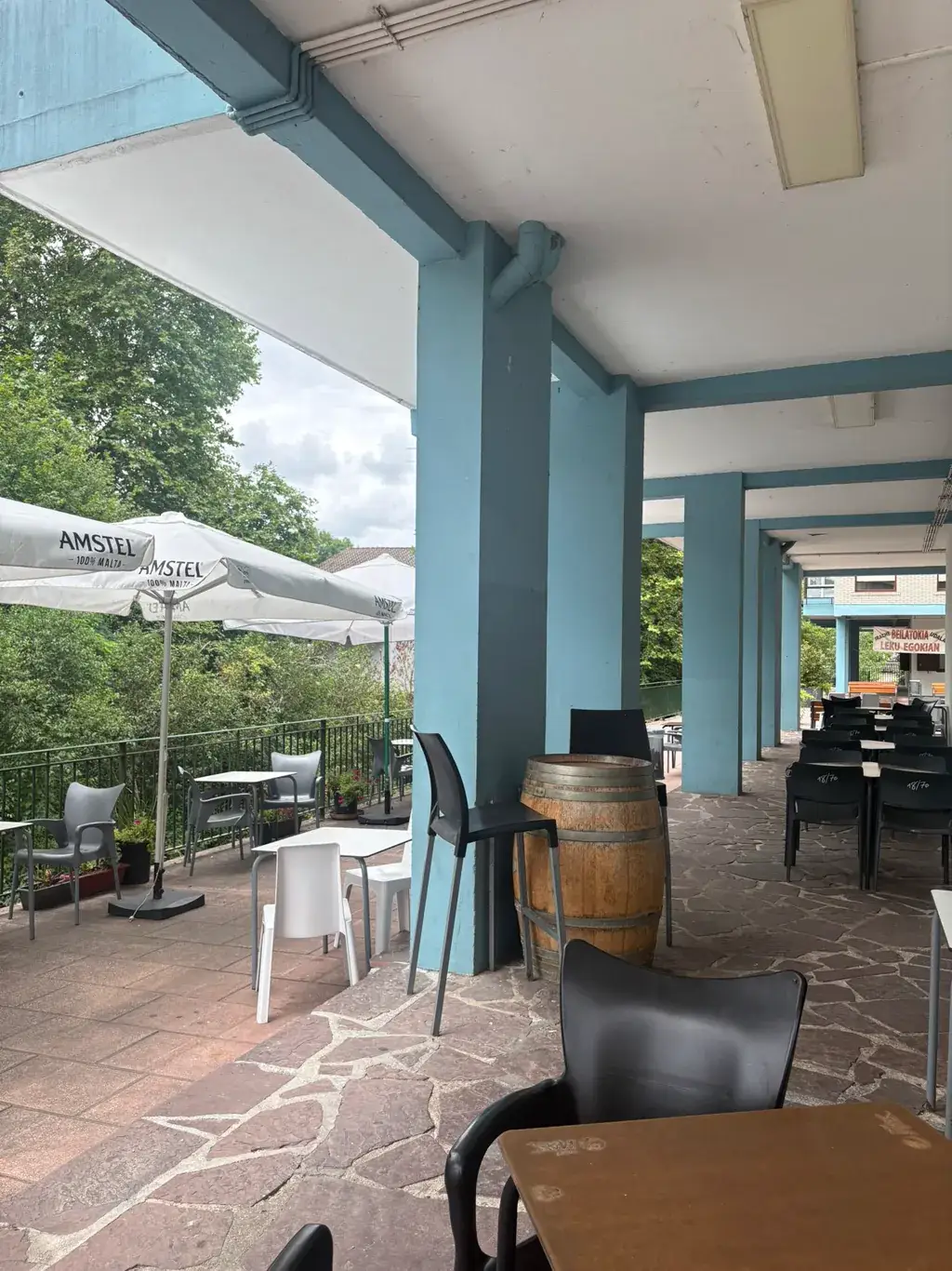 Terraza cubierta de Sara Kafetegia bajo el pórtico de pilares azules, con mesas, sillas y barrica de madera junto a las sombrillas